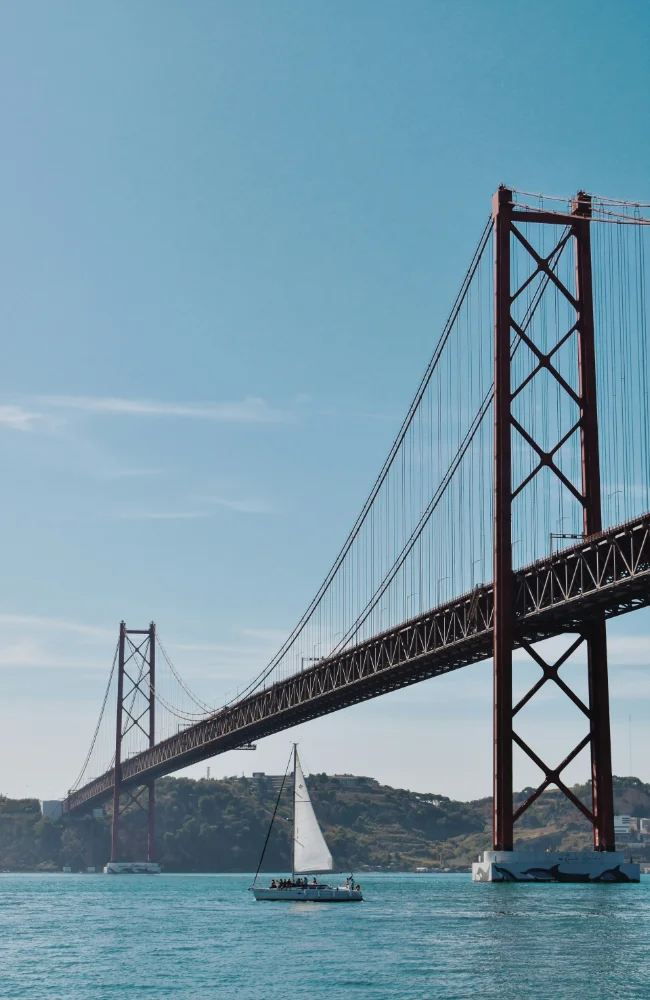 Vue sur un grand pont suspendu de Lisbonne avec un voilier naviguant sur le Tage, montrant le potentiel des événements corporate au Portugal organisés par Solal Events.