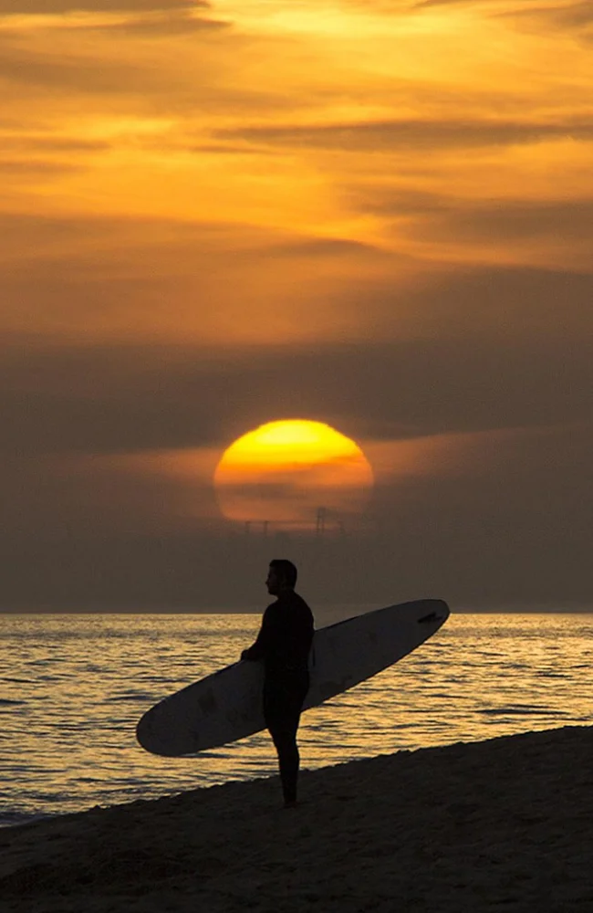 Surfeur portant sa planche face au coucher de soleil sur une plage de l’Algarve, illustrant l’énergie et l’inspiration des événements corporate au Portugal organisés par Solal Events.